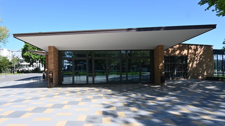 Modern building with glass facade and extended roof, situated on a checker-patterned paved area under a clear blue sky.