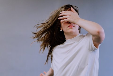 Eine Person mit langen Haaren trägt ein weißes T-Shirt und hält eine Hand vor das Gesicht, während sie sich bewegt.