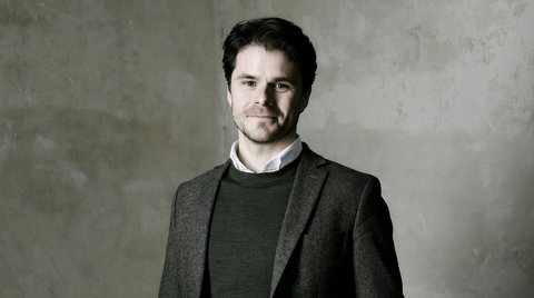 Ein Mann steht in einem dunklen Anzug vor einer grauen, strukturierten Wand. Er schaut direkt in die Kamera.