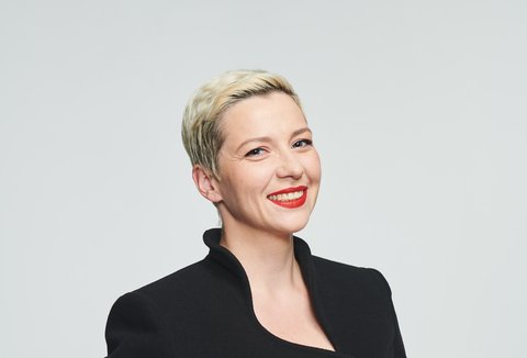 A woman is standing in front of a light-coloured background. She is looking into the camera. Her hair is short and she is wearing a dark jacket. She is smiling.