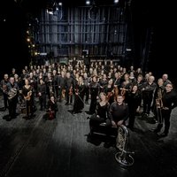 An orchestra is standing in a dark room, turned towards the camera. All are wearing concert clothes and holding instruments.