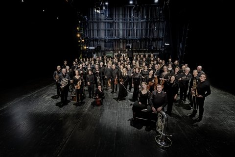 Ein Orchester steht, der Kamera zugewandt, in einem dunklen Raum. Alle tragen Konzertkleidung und halten Instrumente.