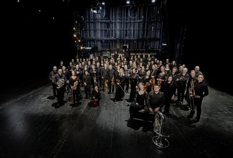 Ein Orchester steht, der Kamera zugewandt, in einem dunklen Raum. Alle tragen Konzertkleidung und halten Instrumente.