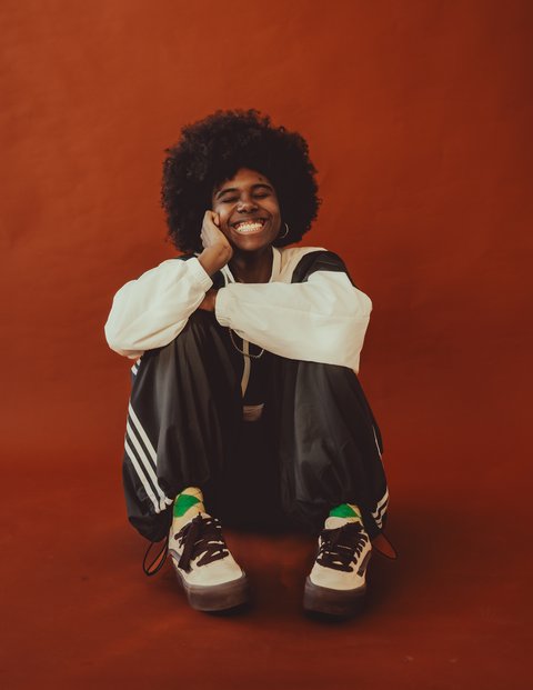 Eine lächelnde Person mit Afro sitzt entspannt mit überkreuzten Beinen auf dem Boden, vor einem orangen Hintergrund.  © Shannon Kone