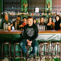 Eine Gruppe von Menschen, das Vokalensemble Roomful of Teeth, steht hinter einer Bar mit verschiedenen Flaschen im Hintergrund. Eine Person, Gabriel Kahane, sitzt auf einem Stuhl vorne.