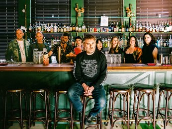 Eine Gruppe von Menschen, das Vokalensemble Roomful of Teeth, steht hinter einer Bar mit verschiedenen Flaschen im Hintergrund. Eine Person, Gabriel Kahane, sitzt auf einem Stuhl vorne.
