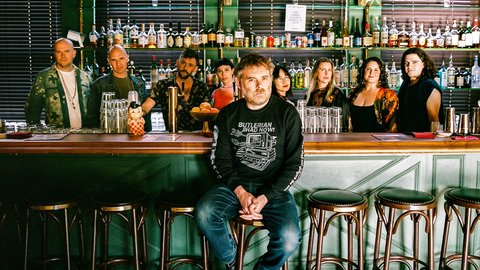 Eine Gruppe von Menschen, das Vokalensemble Roomful of Teeth, steht hinter einer Bar mit verschiedenen Flaschen im Hintergrund. Eine Person, Gabriel Kahane, sitzt auf einem Stuhl vorne. © Charlie Weinmann