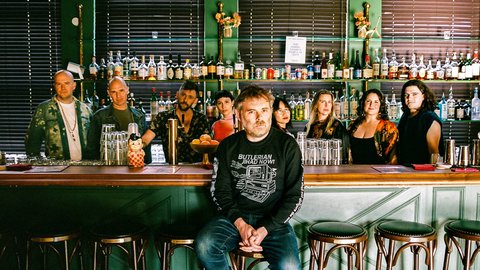 Eine Gruppe von Menschen, das Vokalensemble Roomful of Teeth, steht hinter einer Bar mit verschiedenen Flaschen im Hintergrund. Eine Person, Gabriel Kahane, sitzt auf einem Stuhl vorne.