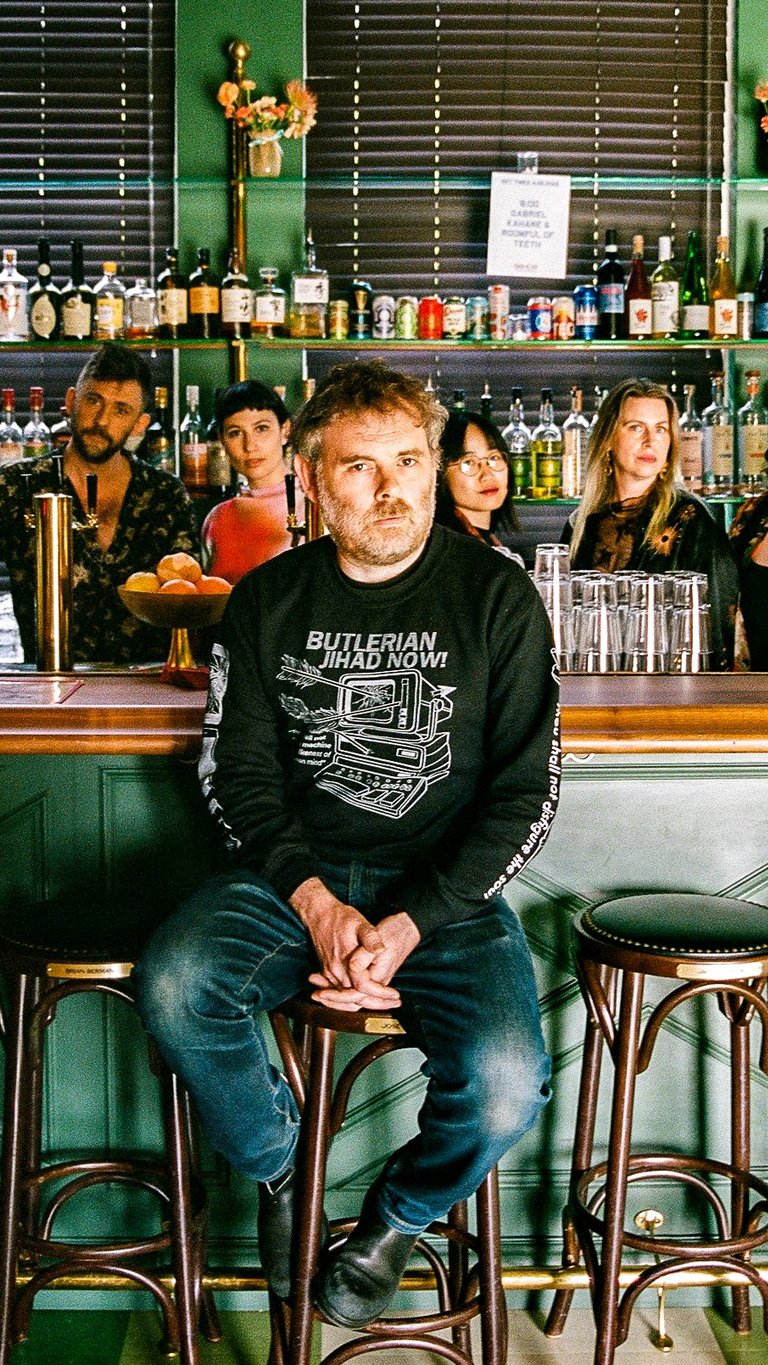 Eine Gruppe von Menschen, das Vokalensemble Roomful of Teeth, steht hinter einer Bar mit verschiedenen Flaschen im Hintergrund. Eine Person, Gabriel Kahane, sitzt auf einem Stuhl vorne.