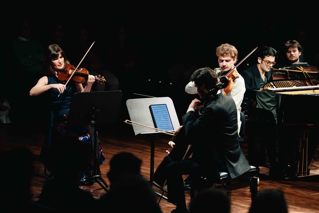 Ein Streichquartett spielt auf einer Bühne, bestehend aus einem Cellisten, Violinisten, Bratschist und Pianisten, im Scheinwerferlicht.