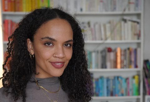 A woman is posing in front of a bookshelf. She has put her hands into her pockets, is looking into the camera and smiling.