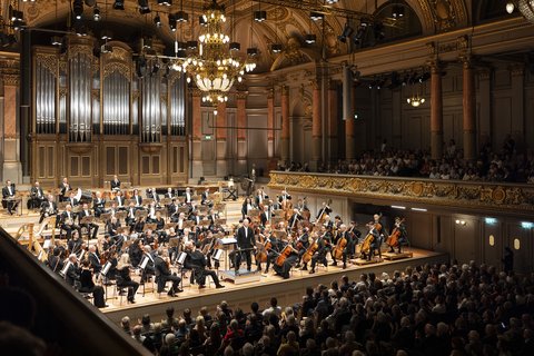 Ein Orchester steht auf der Bühne eines reich geschmückten Saals.