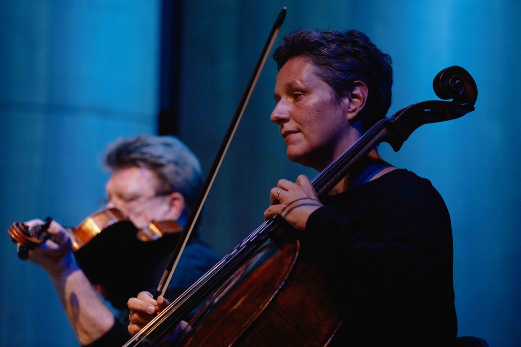 Zwei Musiker auf einer Bühne: ein Cellist im Vordergrund und ein Geiger im Hintergrund, beide konzentriert auf ihr Spiel.