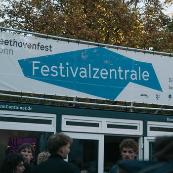 Ein Schild mit der Aufschrift "Festivalzentrale" beim Beethovenfest Bonn. Menschen stehen davor, Bäume im Hintergrund.