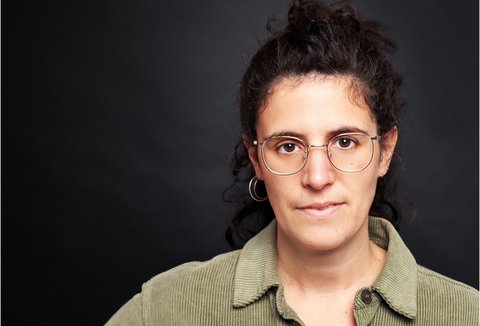 Portrait einer Frau mit Brille und einem grünen Pullover vor einem dunklen Hintergrund. Ihre langen dunklen Haare sind in einen Dutt gefasst und sie blickt in die Kamera.