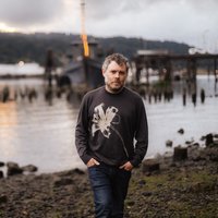 A man stands by a riverside with wooden posts around. The sky is overcast and the atmosphere is dusky.