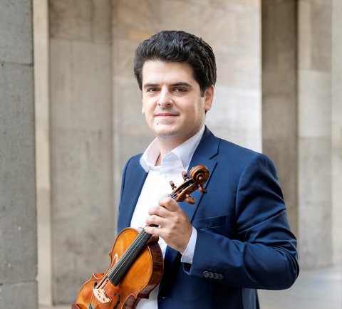 Ein Mann in einem blauen Anzug posiert draußen mit einer Violine in der Hand.