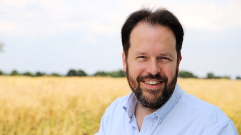 A man is standing before a field in daytime. He is wearing a light-coloured shirt. He is looking into the camera and smiling. © Marshall Light Studio