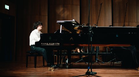 Ein Pianist in einem weißen Hemd spielt auf einer Bühne einen schwarzen Flügel. Der Raum ist dunkel mit Holzwänden.