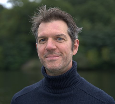 Ein Mann mit dunklem Rollkragenpullover lächelt vor einem Hintergrund aus Bäumen und einem Fluss.