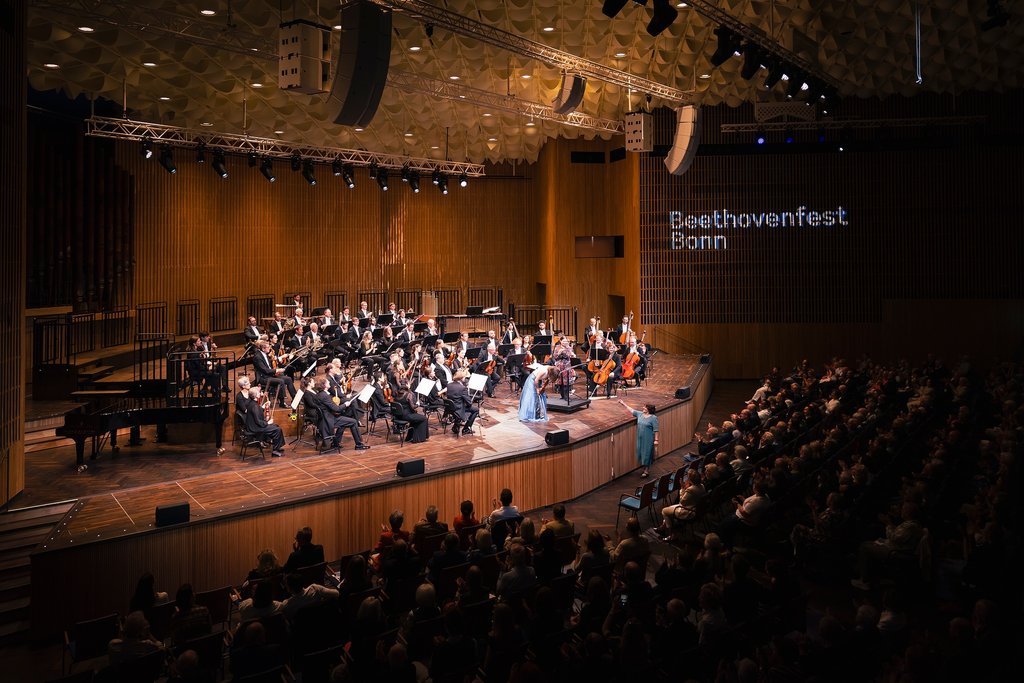 Ein Orchester spielt auf einer großen Bühne. Der Saal ist voll besetzt, und an der Wand steht „Beethovenfest Bonn“.