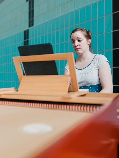 A woman sits at a harpsichord looking at a music stand. The background features blue-green tiled walls.