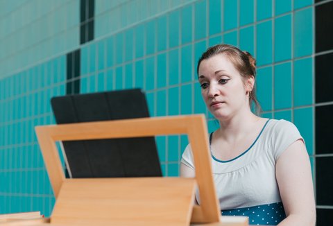 A woman sits at a harpsichord looking at a music stand. The background features blue-green tiled walls.