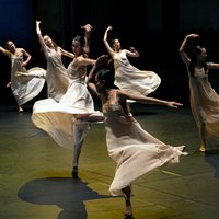 A group of dancers in flowing dresses performs an elegant choreography on a dimly lit stage.