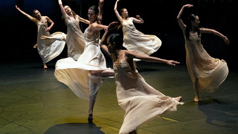 A group of dancers in flowing dresses performs an elegant choreography on a dimly lit stage.