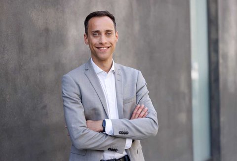 A man in a gray suit stands with crossed arms in front of a gray wall, smiling warmly.