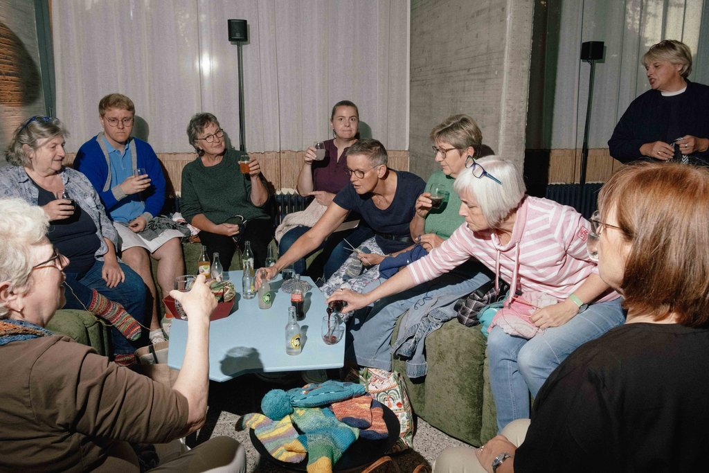 Gruppe älterer Frauen sitzt in einem Raum, trinkt und strickt, umgeben von Wolle und Stricknadeln, in geselliger Atmosphäre.