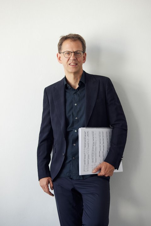 A man in a dark suit stands against a white wall, holding sheet music in his hand.