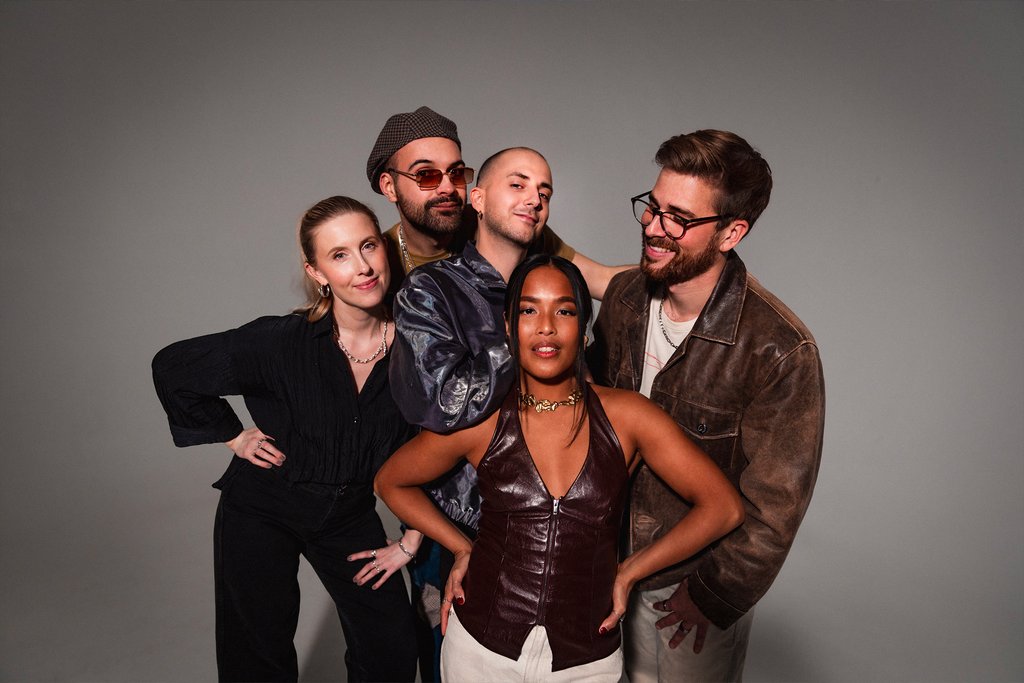 Five people stand together, posing happily. They wear various stylish outfits, exuding a relaxed and friendly atmosphere.