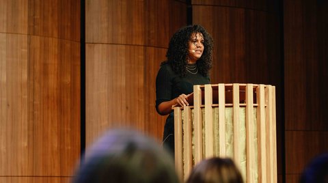 A person speaking at a podium in a wood-paneled room, surrounded by an seated audience.