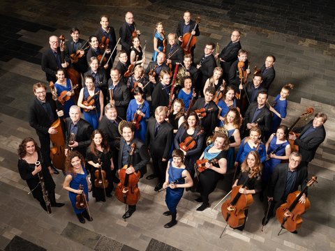 Eine große Gruppe von Orchestermitgliedern in formeller Kleidung posiert mit ihren Streichinstrumenten für ein Gruppenfoto.