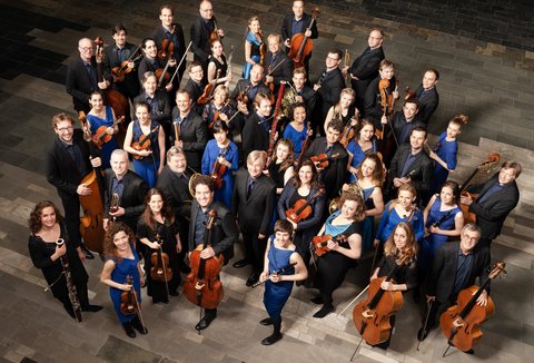 Eine große Gruppe von Orchestermitgliedern in formeller Kleidung posiert mit ihren Streichinstrumenten für ein Gruppenfoto.