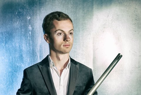 A man is posing in front of a light background, looking into the distance. He is wearing a suit and holding two organ pipes.