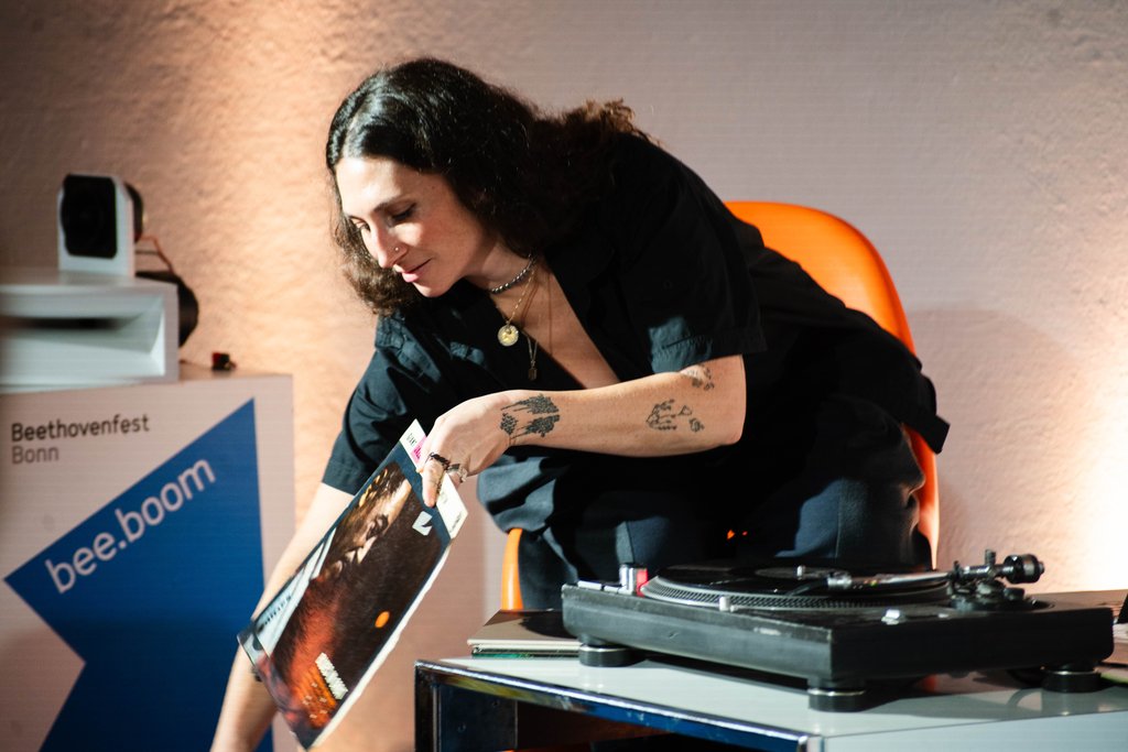 A person with curly hair and tattoos sets up a vinyl record on a turntable during an event.