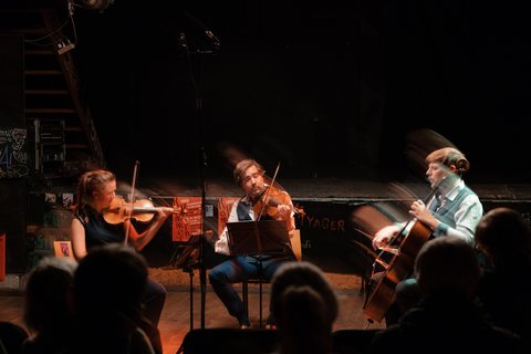Ein Streichtrio spielt auf einer Bühne in einem dunklen Raum, umgeben von einem gespannten Publikum. Personen im Schatten zu erkennen.