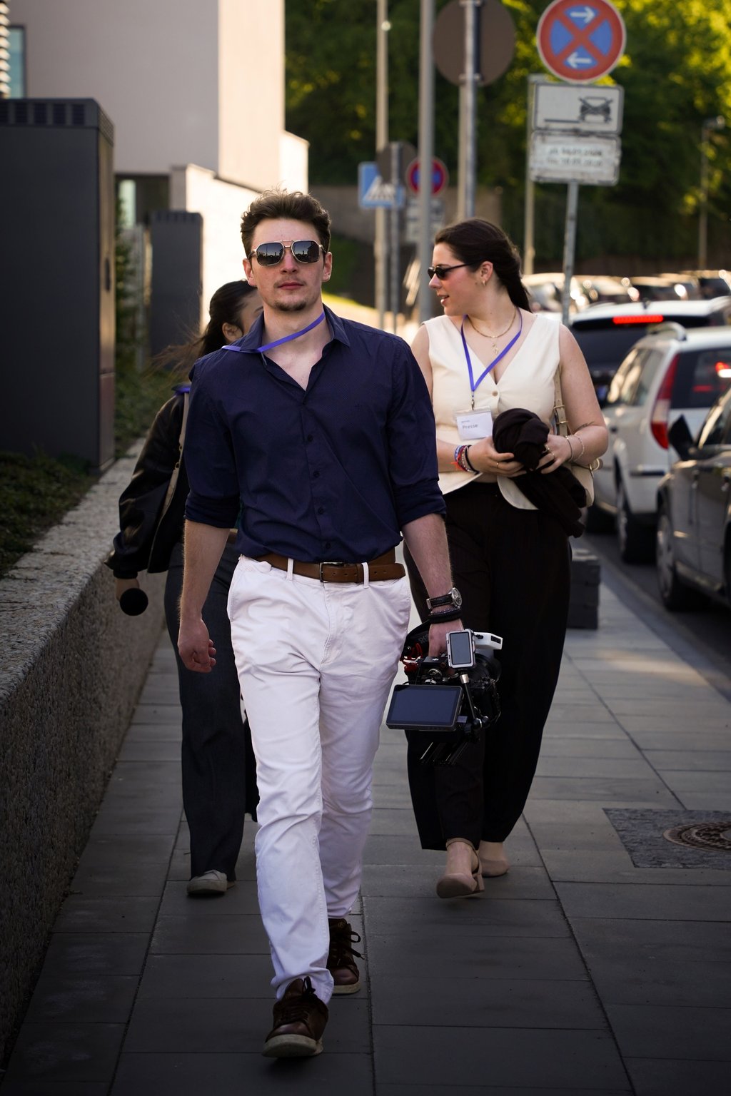 Man in blue shirt and white pants holding a camera, walking on sidewalk with other people in the background.