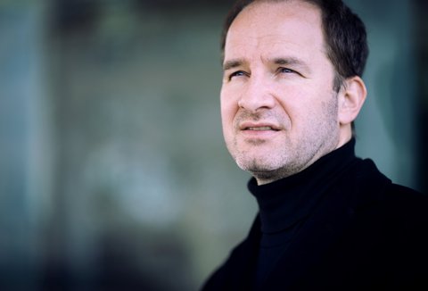 Ein Mann im Porträt, er schaut ernst an der Kamera vorbei. Er trägt einen schwarzen Rollkragenpullover. Der Hintergrund ist türkis-grau.