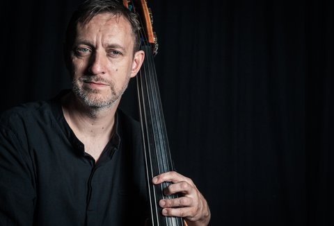 Ein Mann spielt Kontrabass. Er ist gut beleuchtet, während der Hintergrund schwarz ist. Er trägt ein schwarzes Hemd.