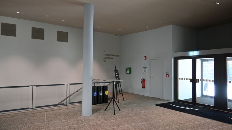 An empty entrance area with a tripod and microphone stand. The walls are white, and daylight streams in from outside.