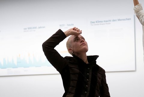 Eine Person in schwarzer Kleidung kniet auf dem Boden in einer Tanzposition. Die Person hat rasierte, blonde Haare und blickt nach rechts oben.