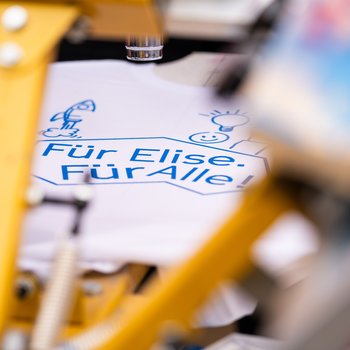 Ein T-Shirt mit der Aufschrift "Für Elise. Für Alle" wird bedruckt. Lebendige Farben und Geräte sind im Vordergrund unscharf.