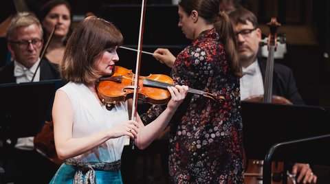 Eine Geigerin spielt leidenschaftlich auf der Bühne, umgeben von Orchestermitgliedern. Die Musiker tragen elegante Konzertkleidung.