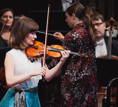 Eine Geigerin spielt leidenschaftlich auf der Bühne, umgeben von Orchestermitgliedern. Die Musiker tragen elegante Konzertkleidung.