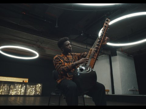 Ein Musiker spielt eine Sitar in einem modernen, dunklen Raum mit kreisförmigen Lichtern. Er trägt ein gemustertes Hemd.