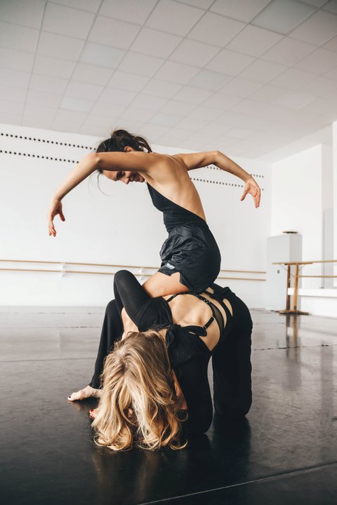 Zwei Tänzer in einem Tanzstudio, der obere balanciert auf dem Rücken des anderen, ihre Körper bilden geschwungene Linien.