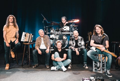Eine Band aus sechs Männern posiert an ihren Instrumenten. Sie blicken in die Kamera und lächeln.